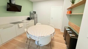 A compact kitchen with a microwave, a coffee maker, and a small refrigerator. There is a round table with a checkered tablecloth and two chairs. A wooden shelf with a plant and a bag is visible. - Bergamoinluxe (Bergamo)