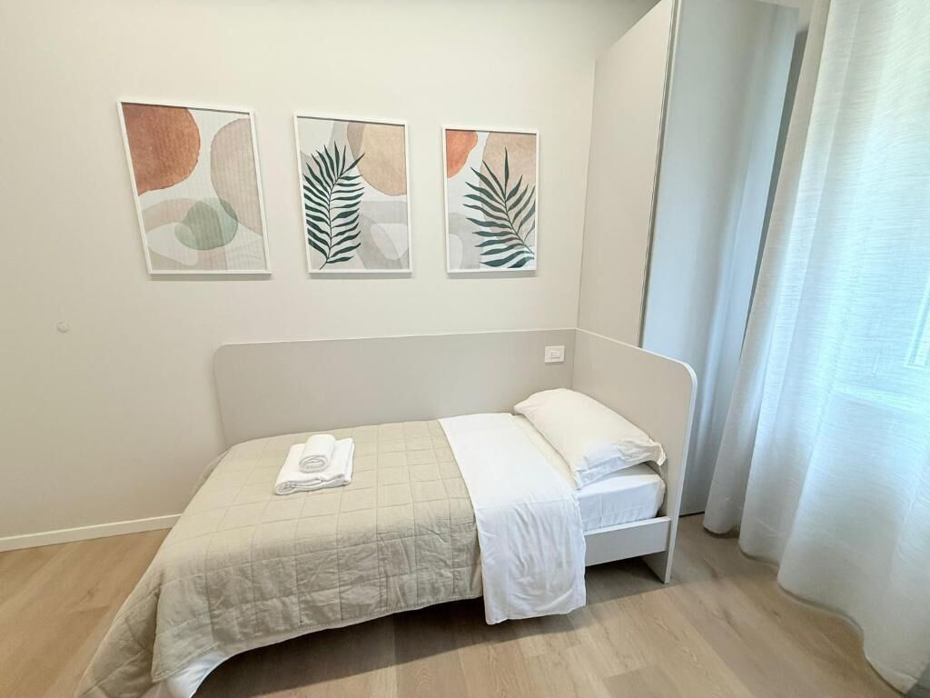 Uma cama de solteiro com colcha branca e toalha dobrada, cabeceira branca e três quadros abstratos de folhas na parede.