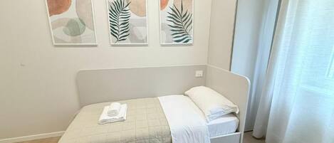 Uma cama de solteiro com colcha branca e toalha dobrada, cabeceira branca e três quadros abstratos de folhas na parede.