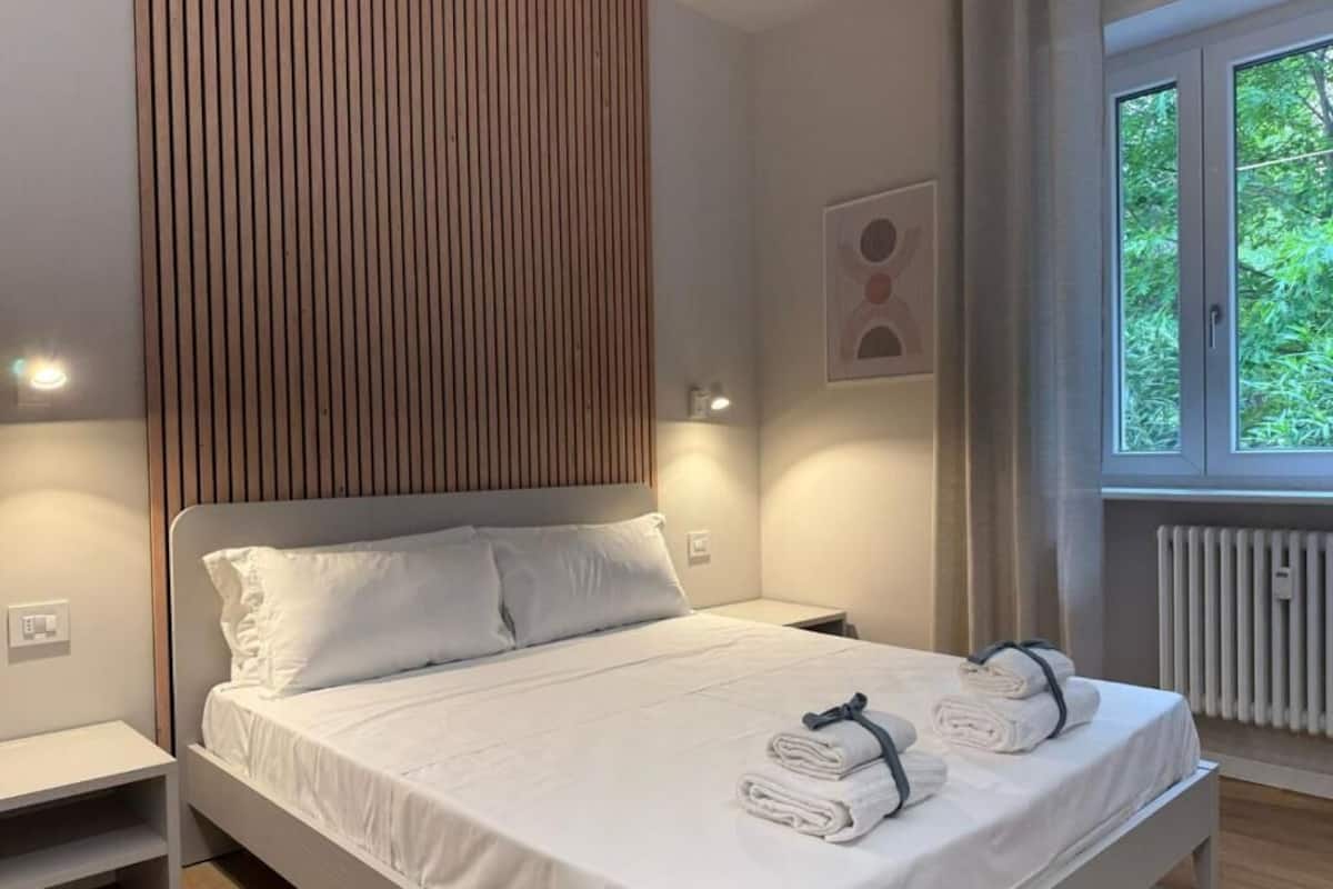 A neatly made bed with white linens and towels, a wooden slat headboard, bedside lamps, and a window with a view of greenery.