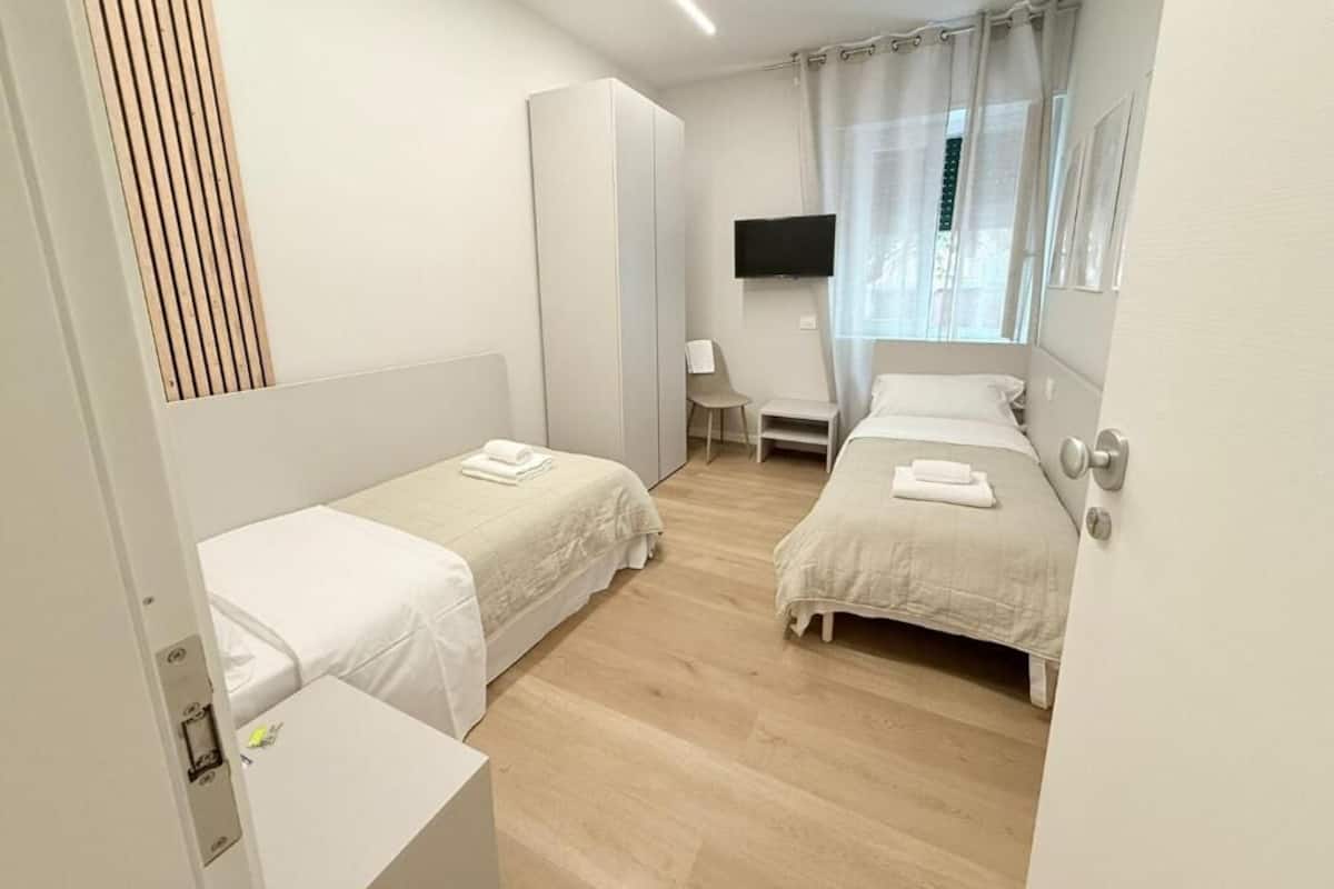 A hotel room with two single beds, a desk, a chair, a TV, and a window with curtains.