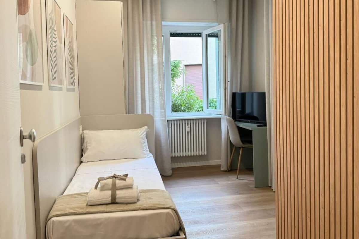 A bedroom with a bed, a desk, a TV, and a window with curtains.