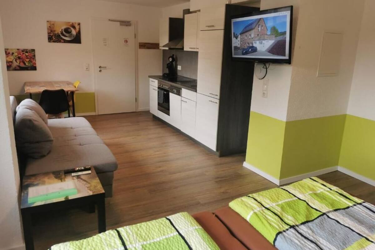 Ein kompakter Wohnbereich mit einer Couch, einem kleinen Esstisch, einer Küchenzeile und einem an der Wand befestigten Fernseher.