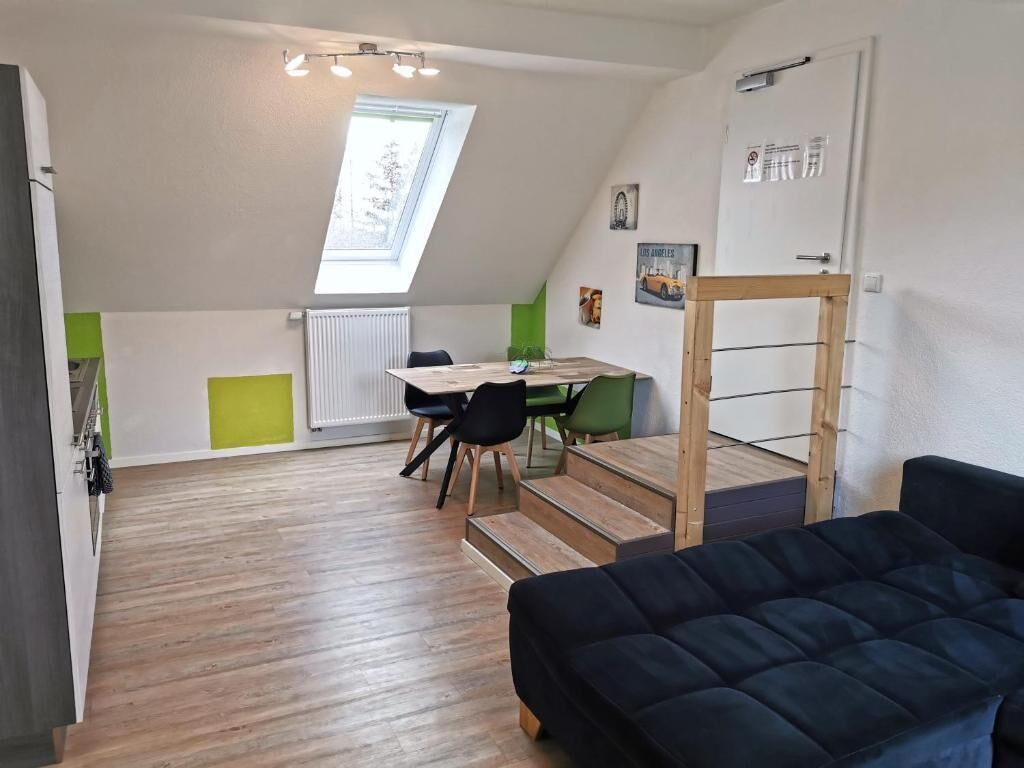 Ein kompakter Wohnraum mit Essbereich, einer schwarzen Couch und einem Schlafzimmer auf Podest mit Fenster.