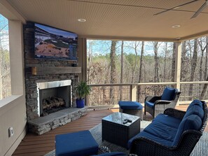 A covered outdoor area with a stone fireplace, a flat-screen TV mounted above it, wicker furniture, and a view of trees. - Masters Week Retreat with Sports Bar, Outdoor Patio & Prime Entertaining Spaces (North Augusta)