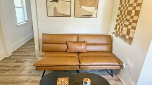 Ein braunes Ledersofa mit Kissen, ein schwarzer Couchtisch mit kariertem Untersetzer und einer kleinen Schale sowie abstrakte Kunst an der Wand.