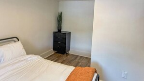 Ein Schlafzimmer mit Bett, Schminktisch, Pflanze und weißer Wand.