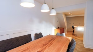 A dining area with a wooden table and chairs, pendant lights, and a view into the kitchen. - Glan y Mor (Porthmadog)