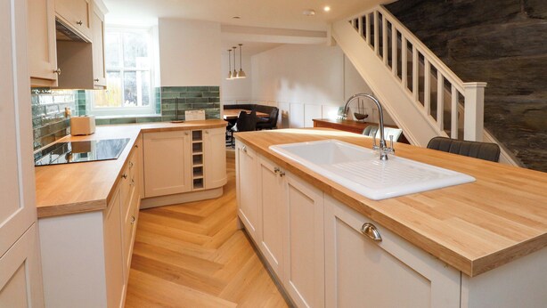 A modern kitchen with wooden cabinetry, a large island with a sink, and a staircase leading to a loft area. - Glan y Mor (Porthmadog)