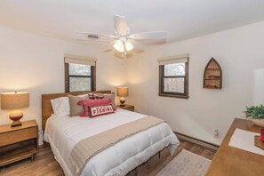 A bedroom with a bed, two bedside lamps, a ceiling fan, and a wooden shelf with decorative items.