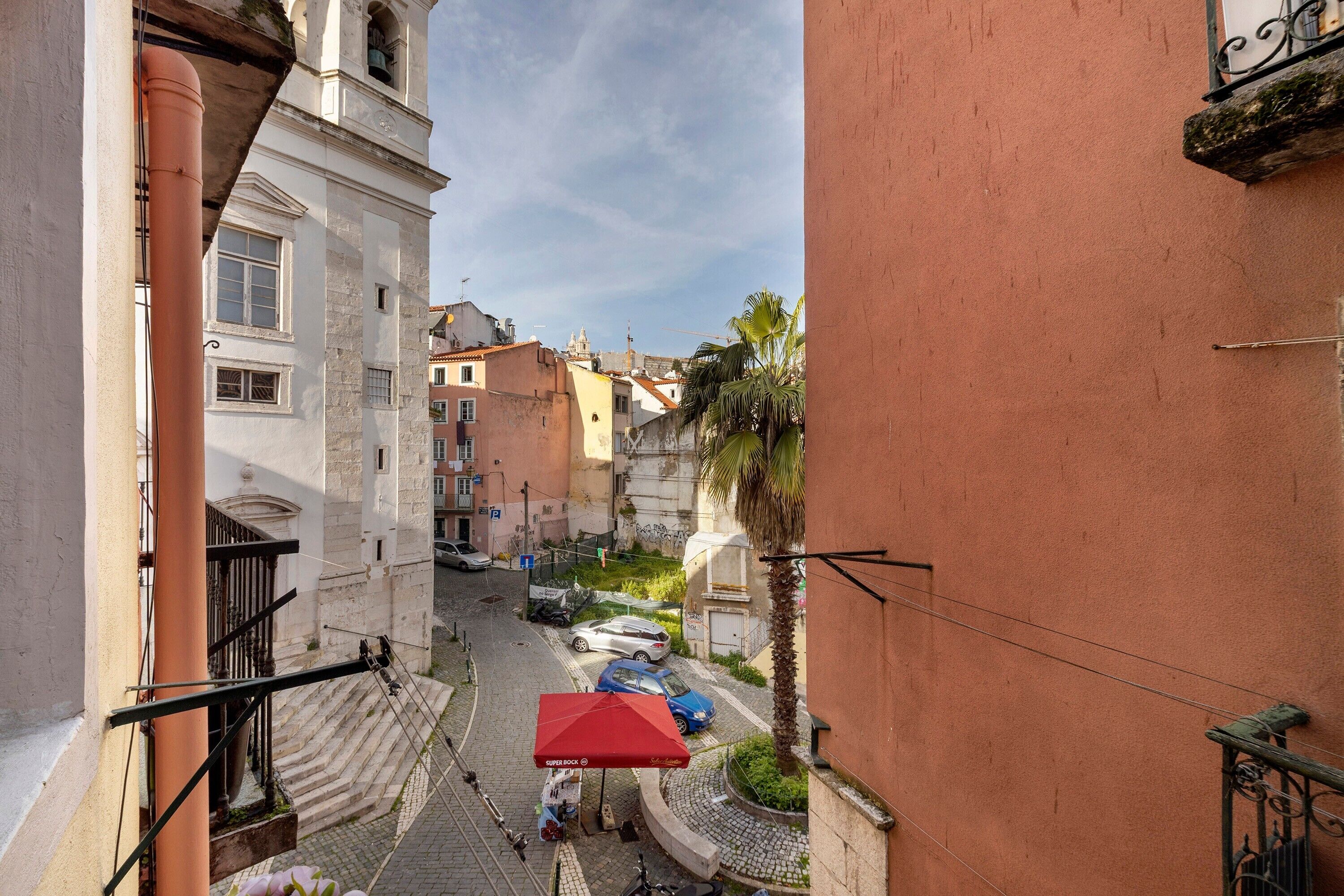 A narrow street with buildings, a red umbrella, and a palm tree.