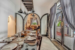 Uma sala de jantar com uma mesa comprida posta para uma refeição, um candelabro e vista através de uma porta arqueada.