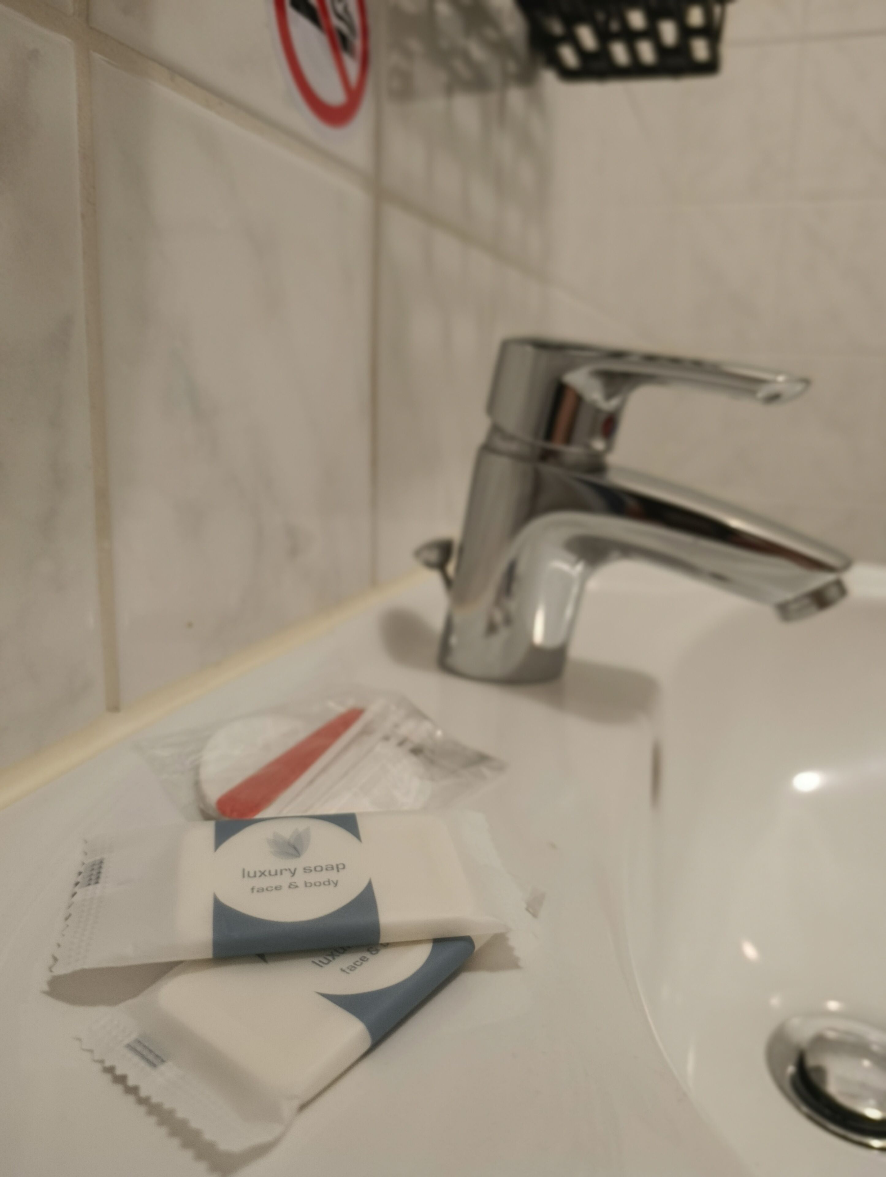 A bathroom sink with a chrome faucet, a no-smoking sign on the wall, and a tube of luxury soap on the counter.