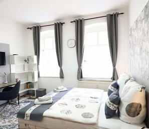 A modern bedroom with a bed, a desk, and a chair. There are two windows with curtains, a clock on the wall, and a radiator. - **MonAppart ** (Gera)