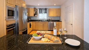 Een moderne keuken met houten kastjes, roestvrijstalen apparatuur, een marmeren aanrechtblad en een ontbijtarrangement met brood, fruit en wijnglazen.