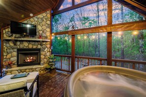 Una chimenea de piedra, un televisor y un jacuzzi en una cabaña con terraza de madera y vistas a árboles.