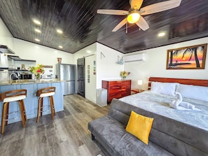 Una habitación de hotel moderna con kitchenette, una cama grande con almohada con forma de cisne, ventilador de techo y una pintura de atardecer.
