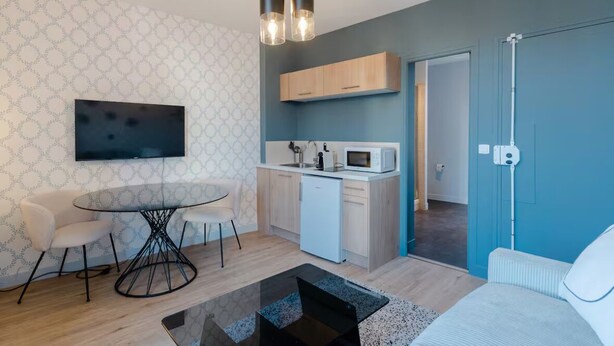 A modern living room with a round table, two chairs, a sofa, a flat-screen TV, a kitchen area with a microwave, and a ceiling light fixture. - Renovated studio with wifi-center of town and beach on foot (Cabourg)