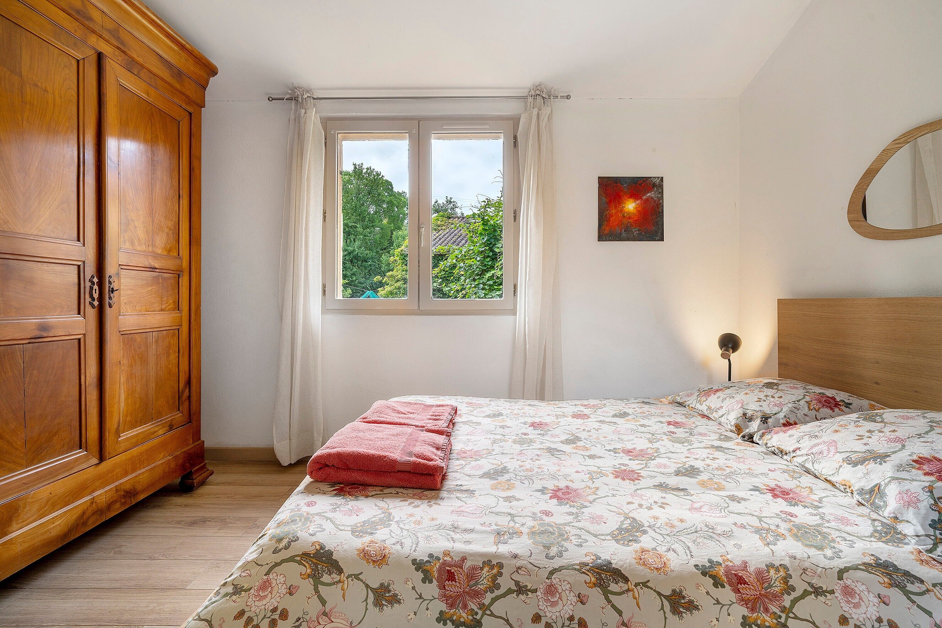 Ein Schlafzimmer mit Holzschrank, einem Bett mit Blumenmuster und einem Fenster mit Vorhängen sowie einem an der Wand befestigten Spiegel.