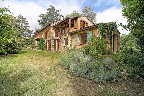 A rustic house with a stone facade, wooden balcony, and lush greenery surrounding the property.