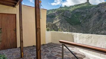 A stone-paved balcony with a wooden railing, a wooden door, and a view of a mountain with a structure on top.