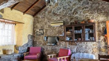 A rustic room with stone walls, wooden beams, a stone fireplace, a television, and a cabinet with dishes.