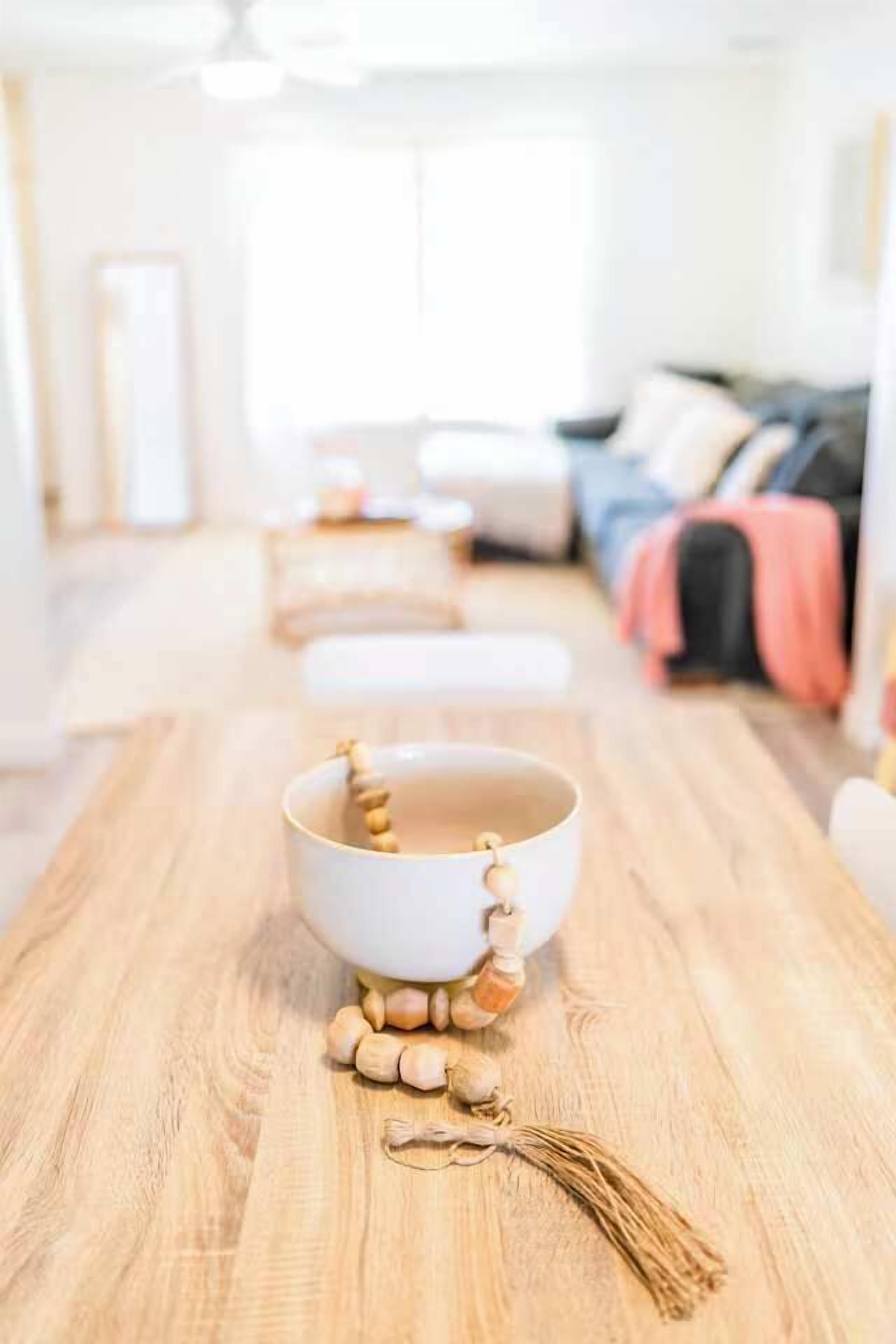 A wooden table with a white bowl and wooden beads, set in a bright room with a sofa and pillows in the background.