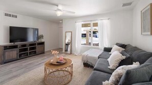 Una sala de estar con un sofá gris, un televisor sobre un mueble de madera, un ventilador de techo y una ventana con cortinas blancas.