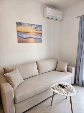 Una sala de estar moderna con un sofá color beige, una mesita redonda con un adorno en la superficie y un cuadro de un atardecer sobre el océano en la pared.