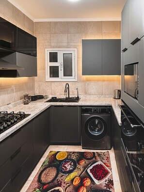 A compact kitchen with dark cabinetry, a built-in oven, a washing machine, and a window above the sink.