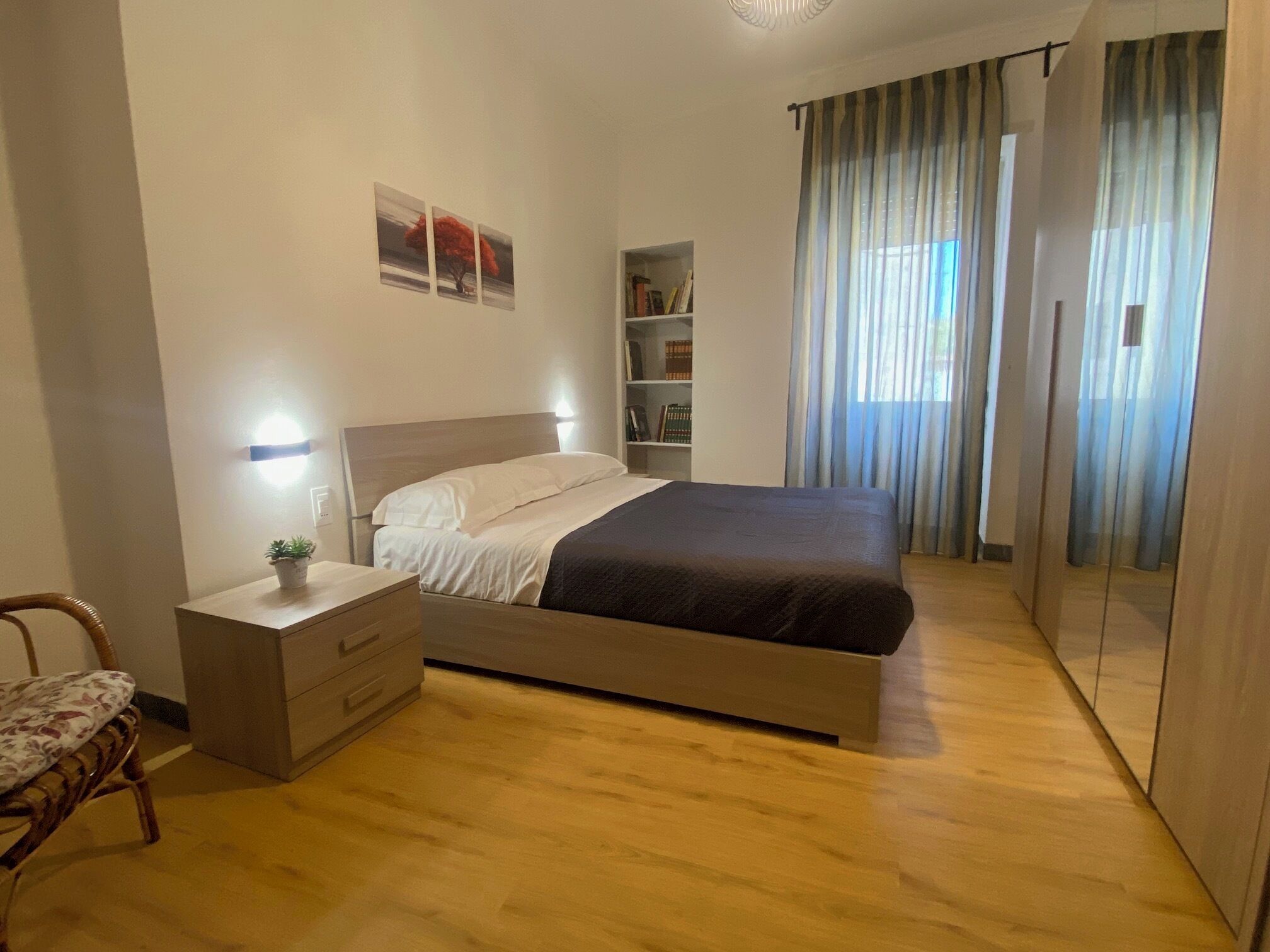 A bedroom with a bed, bedside tables, a chair, a bookshelf, and a window with curtains.