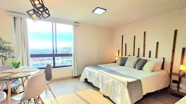 A modern hotel room with a large bed, a dining table, and a view of the city through the window. - New Studio apartment with AC, in front of train station Franklin, Santiago City. (Santiago)