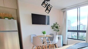 A modern hotel room with a bed, a dining table with chairs, a flat-screen TV, a potted plant, and a large window with a city view. - New Studio apartment with AC, in front of train station Franklin, Santiago City. (Santiago)