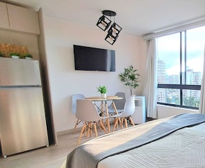 A modern hotel room with a bed, a dining table with chairs, a flat-screen TV, a potted plant, and a large window with a city view. - New Studio apartment with AC, in front of train station Franklin, Santiago City. (Santiago)