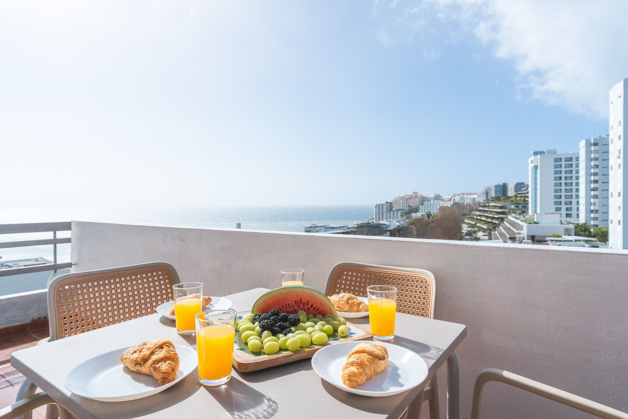 Desayuno en un balcón con vistas a la ciudad, que incluye cruasanes, fruta y jugo de naranja.