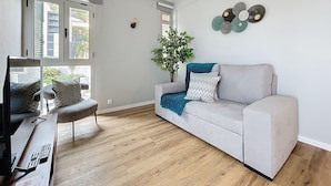 Un salón moderno con un sofá gris, una mesa de centro de madera con controles remotos, una planta en maceta y vistas a la ciudad a través de amplias ventanas.