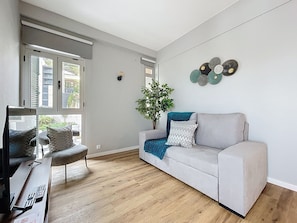 A modern living room with a grey sofa, a wooden coffee table with remote controls, a potted plant, and a city view through large windows. - Lido Edge View by Atlantic Holiday (Funchal)