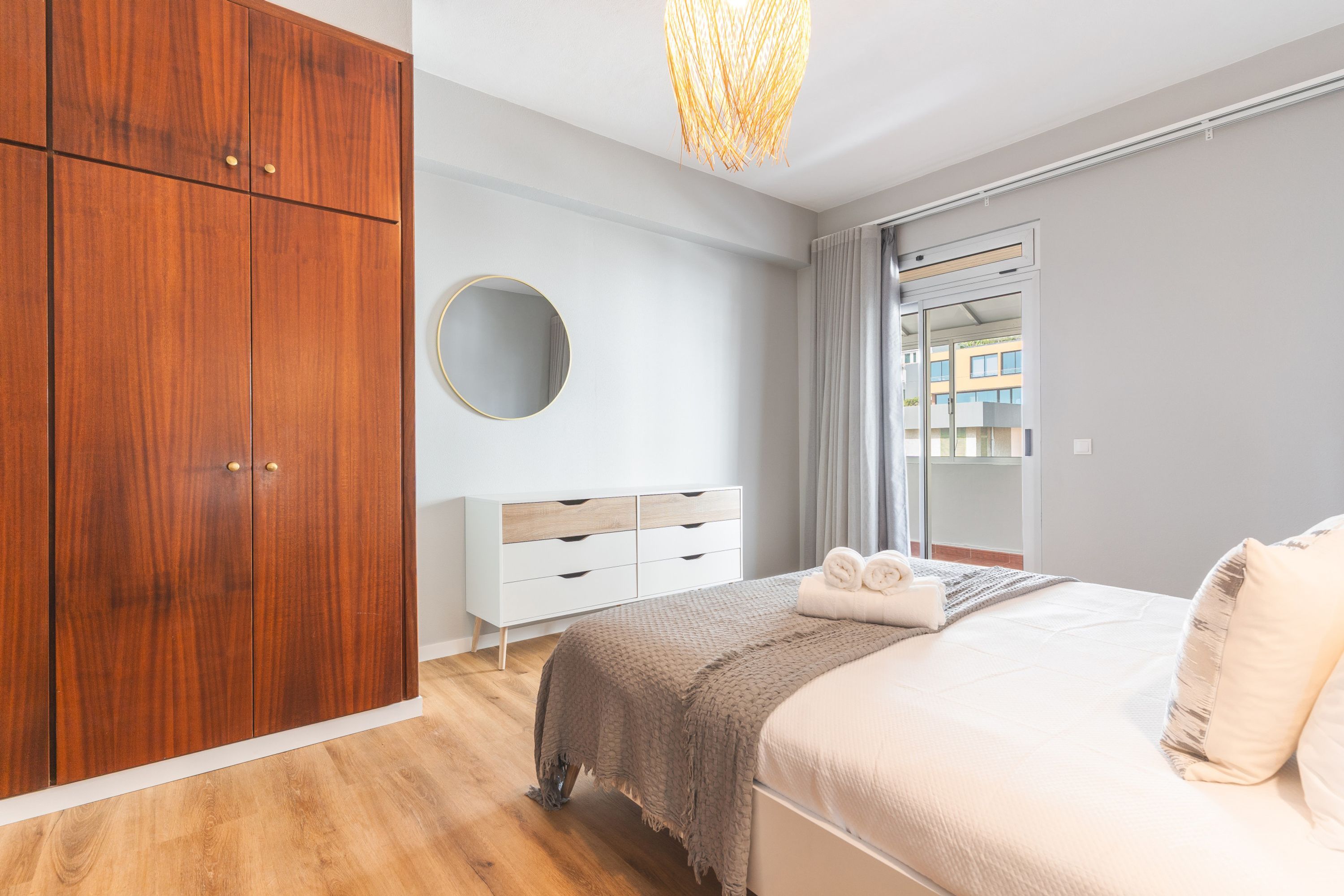 Un dormitorio con un armario de madera, una mesita de noche, un espejo redondo, una cama con una colcha y vistas a través de una ventana.