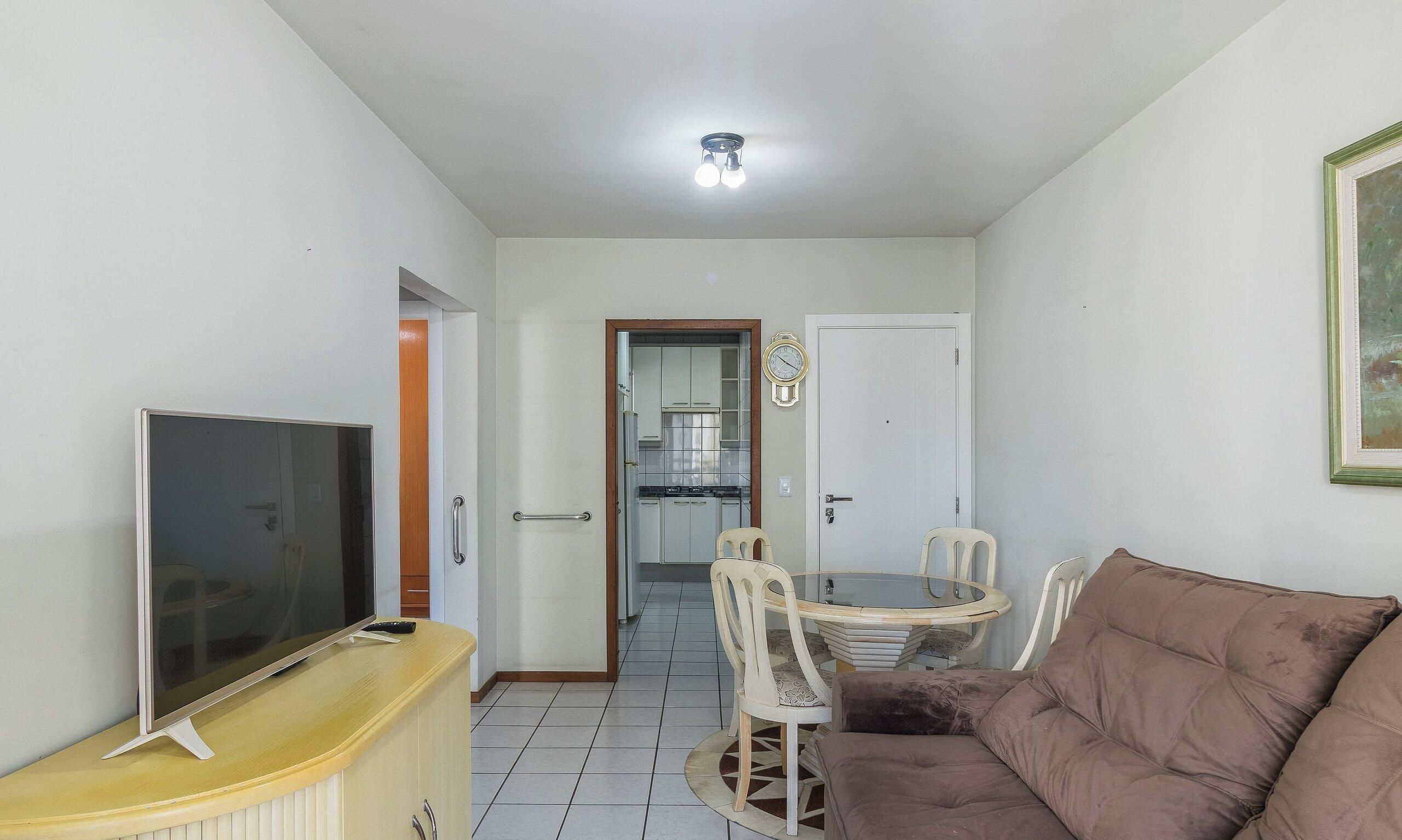 Una sala de estar con televisor, un sofá marrón, una mesa de comedor con sillas y una cocina al fondo.