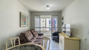 A small, furnished room with a sofa, a dining table, a chair, a TV, and a window with a view of buildings. - Apt in Downtown BC 350m From the Beach Pae1002 (Balneário Camboriú)