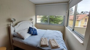 Un lit avec une tête de lit, une fenêtre donnant sur des immeubles et une petite table.