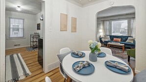 Una sala de estar moderna con un sofá azul, una mesa de comedor con platos y servilletas, y una zona de cocina con ventana y persianas.