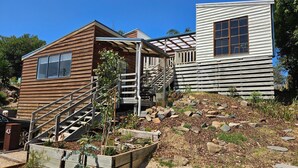 A wooden cabin with a staircase leading to a covered porch, surrounded by a garden and a rocky landscape. - Cedar Hill Retreat - great family property within easy reach of patrolled beach and shops (Venus Bay)