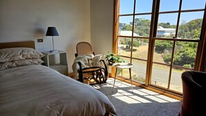 A bedroom with a large window, a bed, a chair, a small table with a plant, and a lamp. - Cedar Hill Retreat - great family property within easy reach of patrolled beach and shops (Venus Bay)