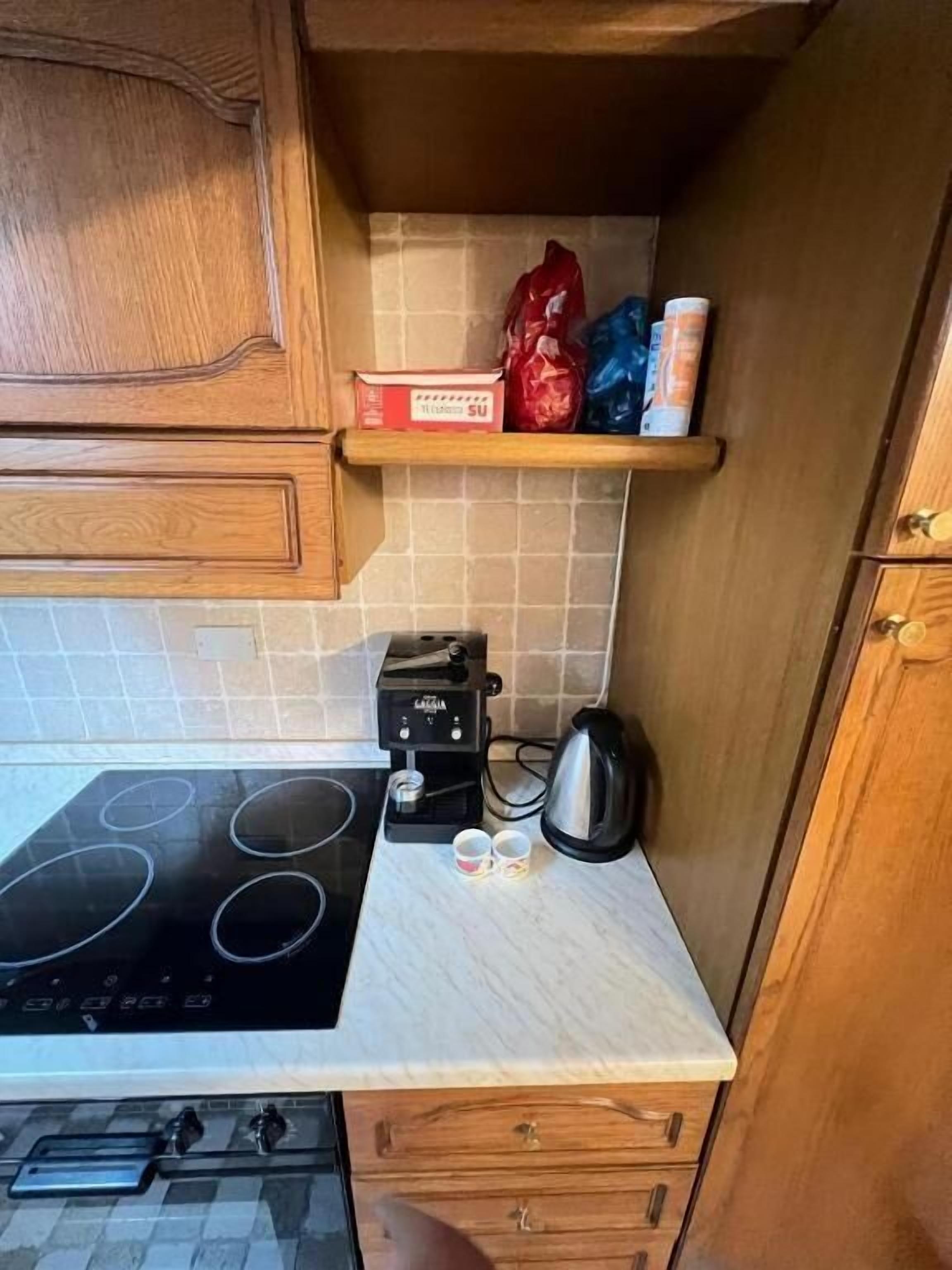 Cocina compacta con estufa negra, cafetera y hervidor en la encimera. Hay armarios de madera y un estante pequeño con algunos objetos.