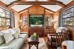 Interior acogedor de una cabaña con techo de madera, un ventanal con vistas a la vegetación, un sofá, una mesa y una silla.