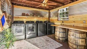 Une buanderie équipée de quatre lave-linge et quatre sèche-linge, avec des poutres en bois, un ventilateur de plafond et une fenêtre donnant sur la forêt.