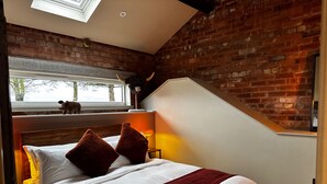 A bedroom with a brick wall, a skylight, a bed with white bedding and brown pillows, a nightstand with a lamp, and a view of a pig through a window. - Pets- Garden- Bbq- Parking- Fireplace- Sleeps 6 (Llanrwst)
