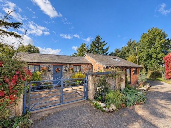 Un bâtiment en pierre avec une porte bleue et une entrée fermée par un portail, entouré d’une végétation luxuriante et d’arbres.