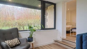 Ett modernt rum med ett stort fönster, en grå fåtölj med en dekorativ kudde, ett litet bord med en krukväxt och en säng med ett färgglatt överkast.